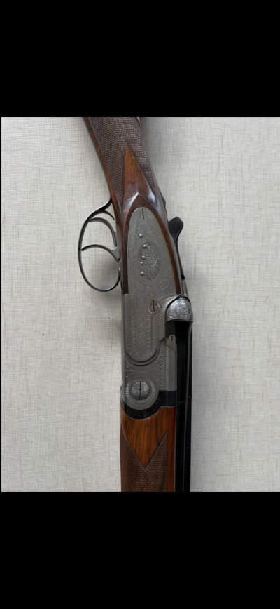 Espingarda de caça de madeira e metal com gravações ornamentais e gatilho duplo num fundo cinzento liso.
