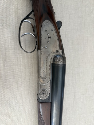 Espingarda com coronha de madeira escura e detalhes em metal prateado gravado, apoiada em fundo cinza claro