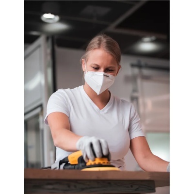 Mulher lixa madeira com lixadora elétrica usando máscara e luvas.