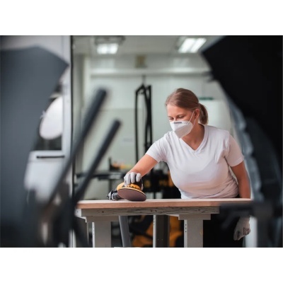 Mulher a lixar madeira numa oficina com lixadeira amarela e preta.