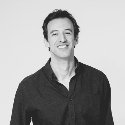 Homem a sorrir com camisa de botões preta em fundo branco preto e branco