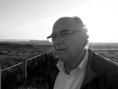 Homem de meia-idade com óculos e casaco ao ar livre junto a uma cerca em área aberta.