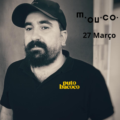 Homem com boné preto e t-shirt preta com texto amarelo puto bacoco em fundo cinza