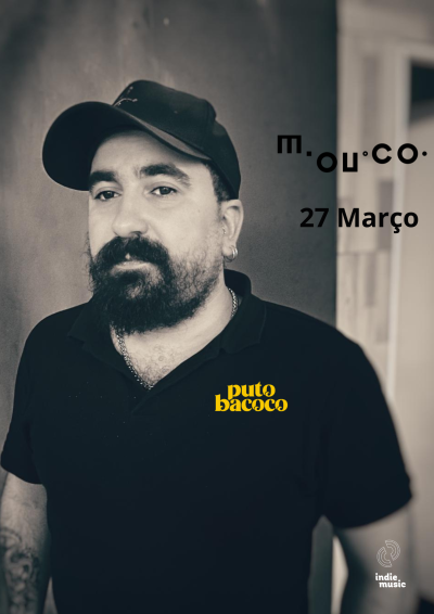 Homem com boné preto e t-shirt preta com texto amarelo puto bacoco em fundo cinza