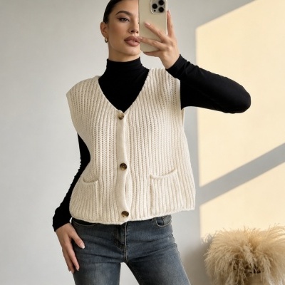 Colete feminino bege claro de malha com botões e bolsos, usado sobre camisa preta e jeans azuis