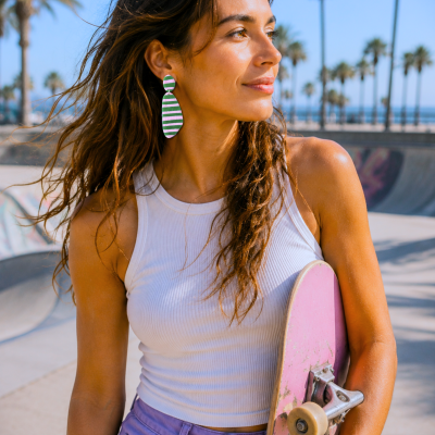 Brincos grandes com riscas rosa e verdes usados por mulher com top branco e calças roxas segurando skateboard rosa ao ar livre.