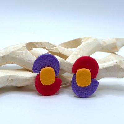 Brincos coloridos vermelho, roxo e amarelo com fundo branco e ornamento de madeira.