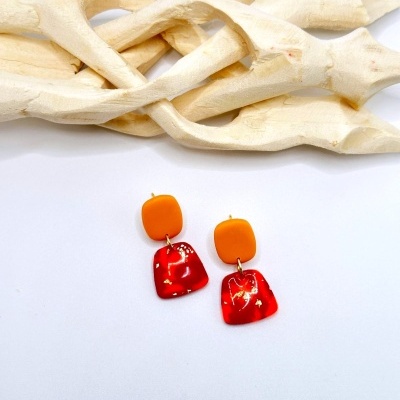 Par de brincos laranja e vermelho com detalhes dourados sobre fundo branco e madeira clara