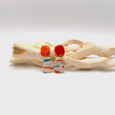 Brincos pendentes laranja e branco com padrão azul e laranja sobre fundo branco e madeira clara.