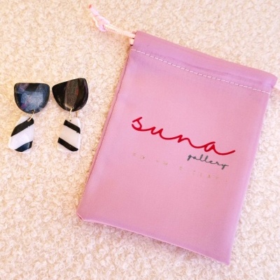 Brincos bicolores ao lado de bolsa de tecido rosa com logotipo