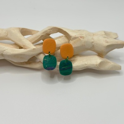 Brincos laranja e verde com textura detalhada, sobre estrutura de madeira clara.