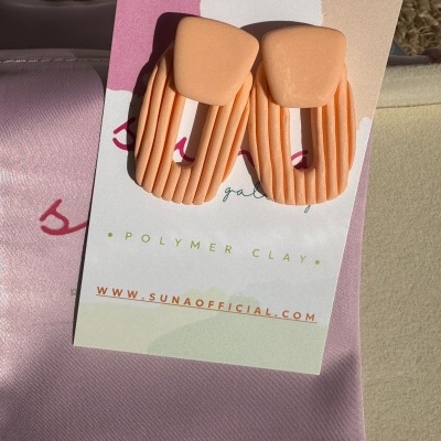 Brincos cor de pêssego de argila de polímero sobre cartão com texto, sobre tecido rosa claro.