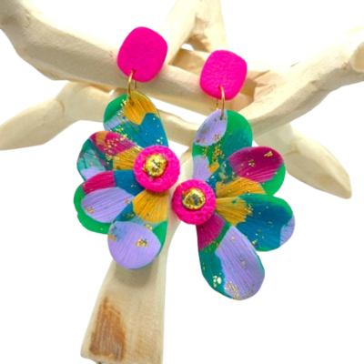 Brincos coloridos em forma de flor com base rosa pendurados numa estrutura branca.