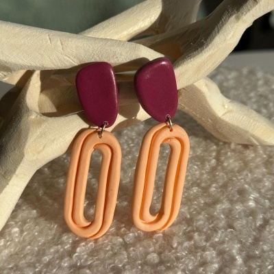 Brincos em cores vinho e pêssego pendurados numa escultura de madeira branca