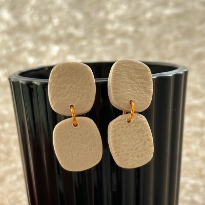 Brincos bege em duas peças quadradas com argolas douradas, sobre copo preto canelado