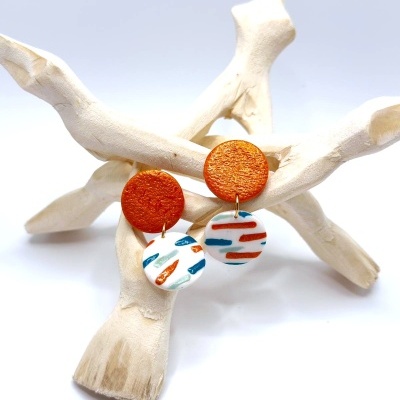 Brincos com discos laranja e branco com padrões coloridos em suporte de galho de madeira branco.