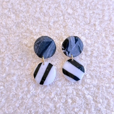 Brincos de argila cinzenta e branca com riscas pretas sobre tecido branco texturado.