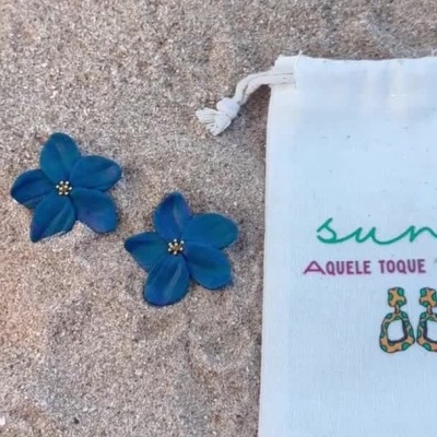Brincos flor azul na areia ao lado de saco branco com texto