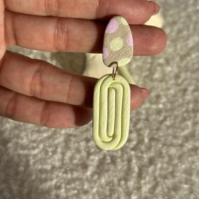 Brinco pendente colorido nas cores bege, rosa, amarelo segurado por mão em fundo branco