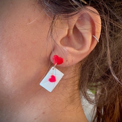 Brinco com envelope branco e corações vermelhos na orelha de uma pessoa