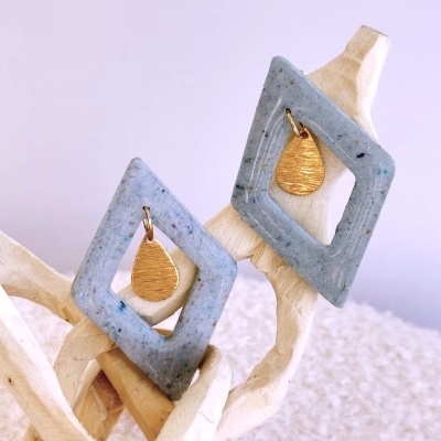 Brincos azuis cinzentos geométricos com pingente dourado em forma de gota sobre galho de madeira clara