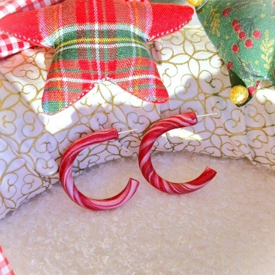 Brincos em argola vermelhos e brancos com padrão torcido sobre superfície branca e almofadas natalícias ao fundo.