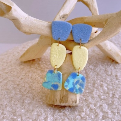 Brincos pendentes tricolor azul e amarelo com textura mate sobre ramo de madeira clara