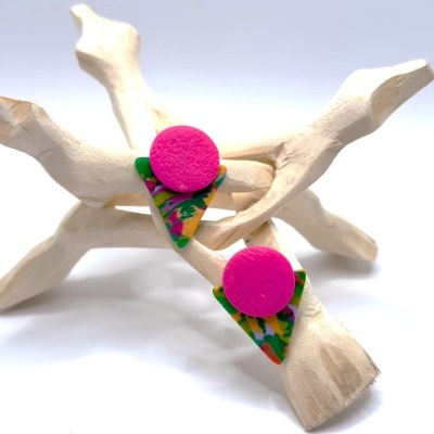 Brincos coloridos de acrílico em formas de círculo rosa e triângulo multicolorido sobre galhos secos brancos.