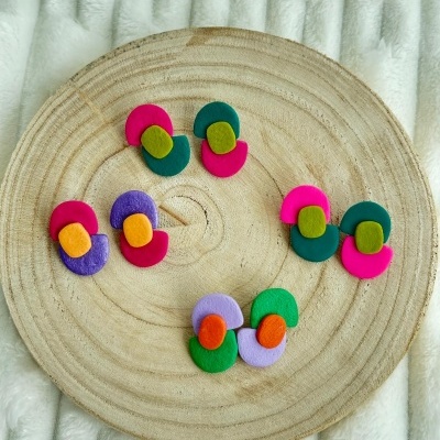 Conjunto de brincos coloridos em madeira, dispostos em placa circular com fundo de tecido branco.