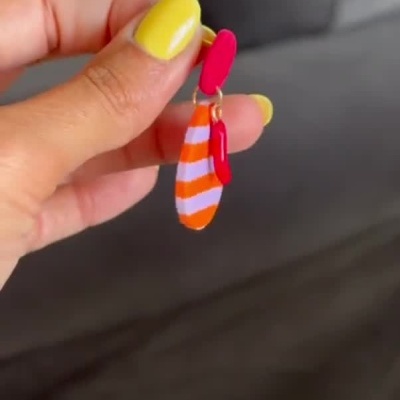 Brinco colorido laranja com riscas lilases e pendente vermelho segurado por mão com unhas amarelas