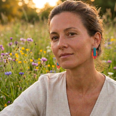 Mulher com brincos coloridos em azul, vermelho e laranja, em campo florido ao pôr do sol.