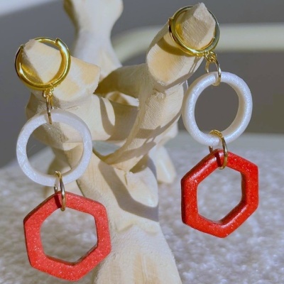 Brincos dourados com argolas branca e vermelha pendurados num suporte de madeira