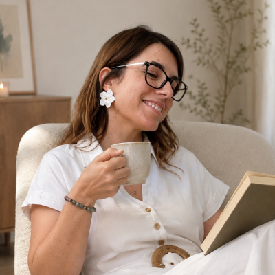 Mulher com vestido branco e brincos flor branca segurando caneca e livro em poltrona creme