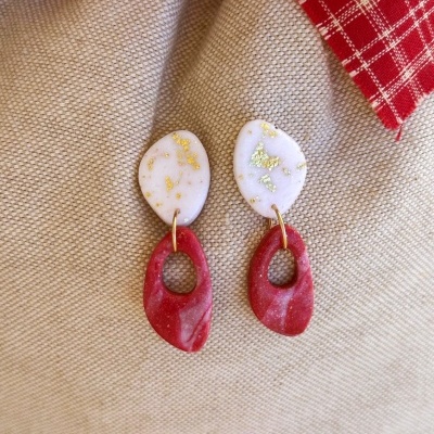 Brincos brancos com detalhes dourados e vermelhos marmoreados sobre fundo bege com tecido vermelho xadrez