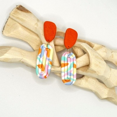 Brincos coloridos em resina com padrão multicolor sobre fundo branco e madeira clara.