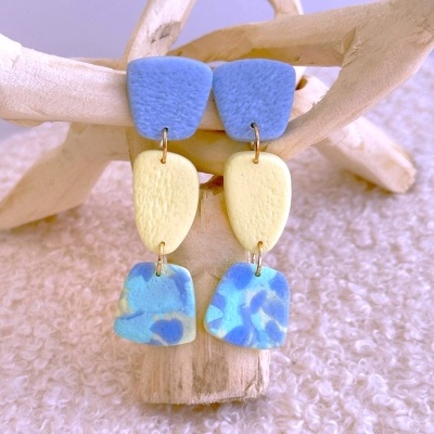 Brincos pendentes azul e amarelo com textura sobre fundo branco e estrutura de madeira