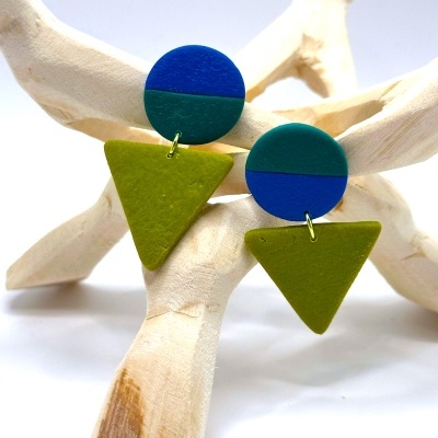 Brincos geométricos azul e verde pendurados em estrutura de madeira branca.