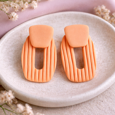 Par de brincos laranja ovais com linhas verticais em placa branca com flores brancas