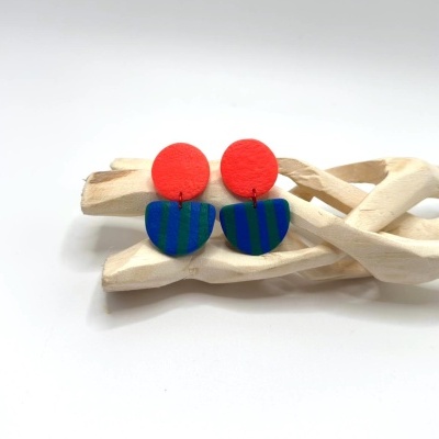 Brincos geométricos vermelho e azul com riscas verdes sobre madeira clara entrelaçada