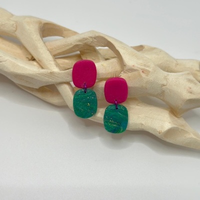 Brincos de resina magenta e azul esverdeado com padrão marmoreado sobre estrutura de madeira clara.