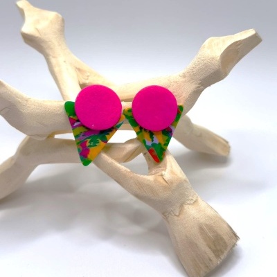 Brincos coloridos com base redonda rosa e triângulo multicolorido sobre suporte de madeira clara.