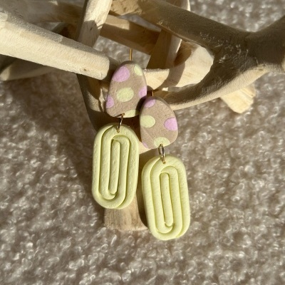 Brincos pendentes em argila polimérica com padrão de bolinhas e formas ovais amarelas, expostos sobre base de madeira e tecido fofo.