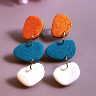 Brincos pendentes coloridos em laranja, azul e branco sobre fundo rosa.