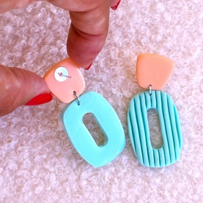 Brincos coloridos em pastel com formas geométricas sobre superfície branca texturada.