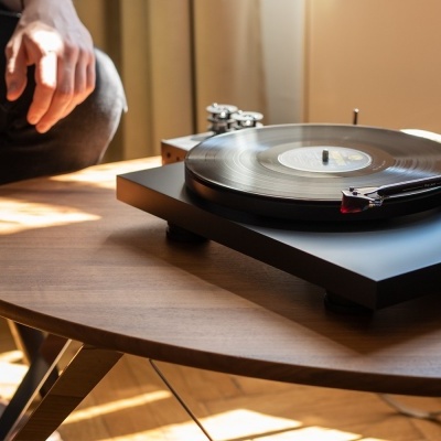 Gira-discos preto a tocar num vinil numa mesa de madeira clara junto a uma pessoa sentada.