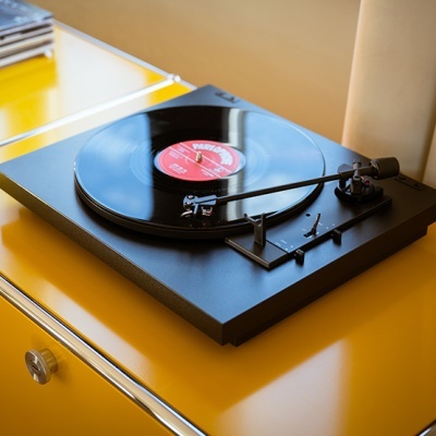 Toca-discos preto com disco de vinil e etiqueta vermelha sobre móvel amarelo