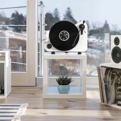 Gira-discos branco com disco preto numa mesa branca com planta, estante com discos e altifalante numa sala moderna com piso de madeira e vista para neve.