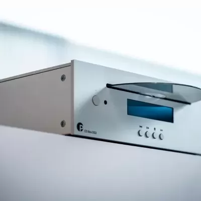 Aparelho eletrónico prateado com visor digital azul e botões, sobre superfície branca