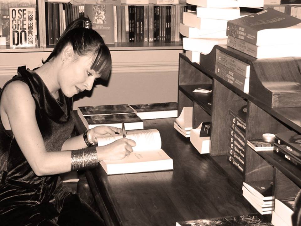 Mulher a escrever num livro numa mesa em biblioteca com livros ao fundo