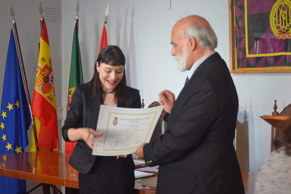 Sessão Solene de Tomada de Posse: Academia de Letras e Artes - Portugal Homem entrega diploma a mulher sorridente em sala com bandeiras e mesa de madeira.